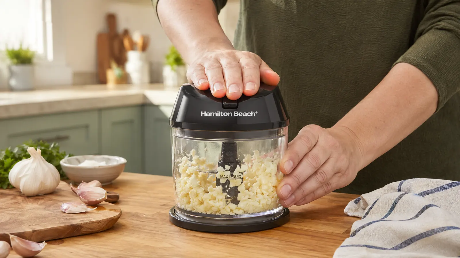 Hand pressing the lid down on the Hamilton Beach food chopper to pulse-chop a batch of garlic cloves in the clear bowl